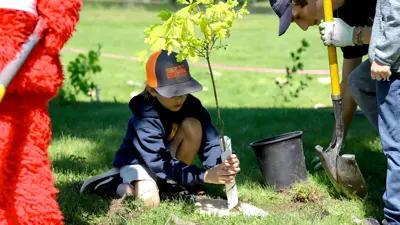 Two individuals and a mascot planting a tree outdoors.