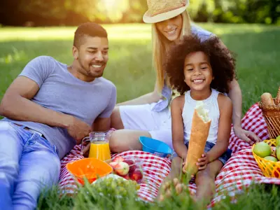 A joyful family enjoying a picnic in a sunny park, with bright smiles, a spread of fruits and snacks on a red blanket.