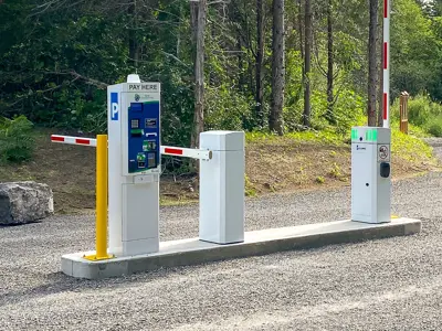 Entrance to a parking area with an automated payment machine labeled "PAY HERE" and a barrier gate, set against a background of trees.