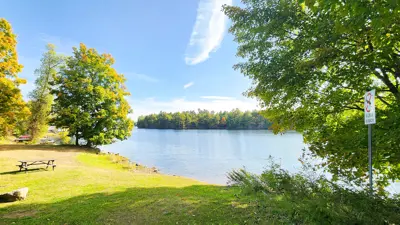 View of a tranquil lake surrounded by autumn-colored trees under a clear sky. A sign that reads "No Swimming" is visible on the right, alongside a picnic area with tables and rocks.