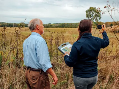 Two people are observing a natural landscape, one holding a map and the other taking a photo with a smartphone.