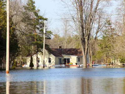 A residential area experiencing significant flooding with water reaching the walls of homes and covering the street. Several trees and utility poles are also visible in the submerged area.
