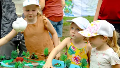 Three children are intently observing a model display at an outdoor event, with one child pointing at a detail on the model.