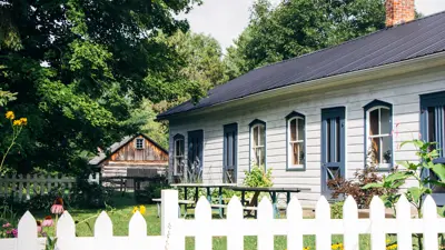 A quaint countryside cottage with a white picket fence surrounded by a lush garden, under a clear sky.