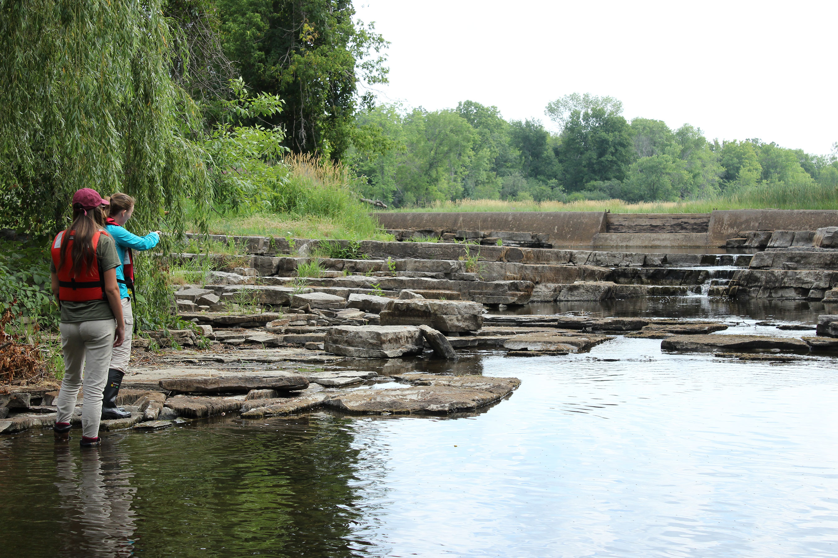 Two people stand by a rocky stream, pointing towards a dam.