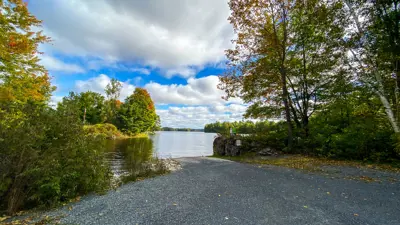 Scenic view of a lake bordered by trees displaying early autumn colors under a partly cloudy sky. A gravel path leads towards the water.