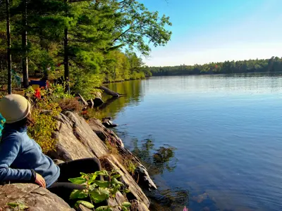 A person sitting on a rocky lakeside, gazing at the serene waters and forest under a clear blue sky.