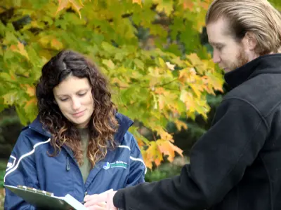 Two individuals in an outdoor setting, one wearing a jacket with the logo "Quinte Conservation," are looking at a clipboard together.