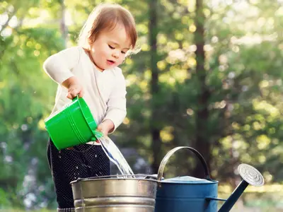 A child pours water from a green watering can into a large metal bucket outdoors, surrounded by sunlit trees.