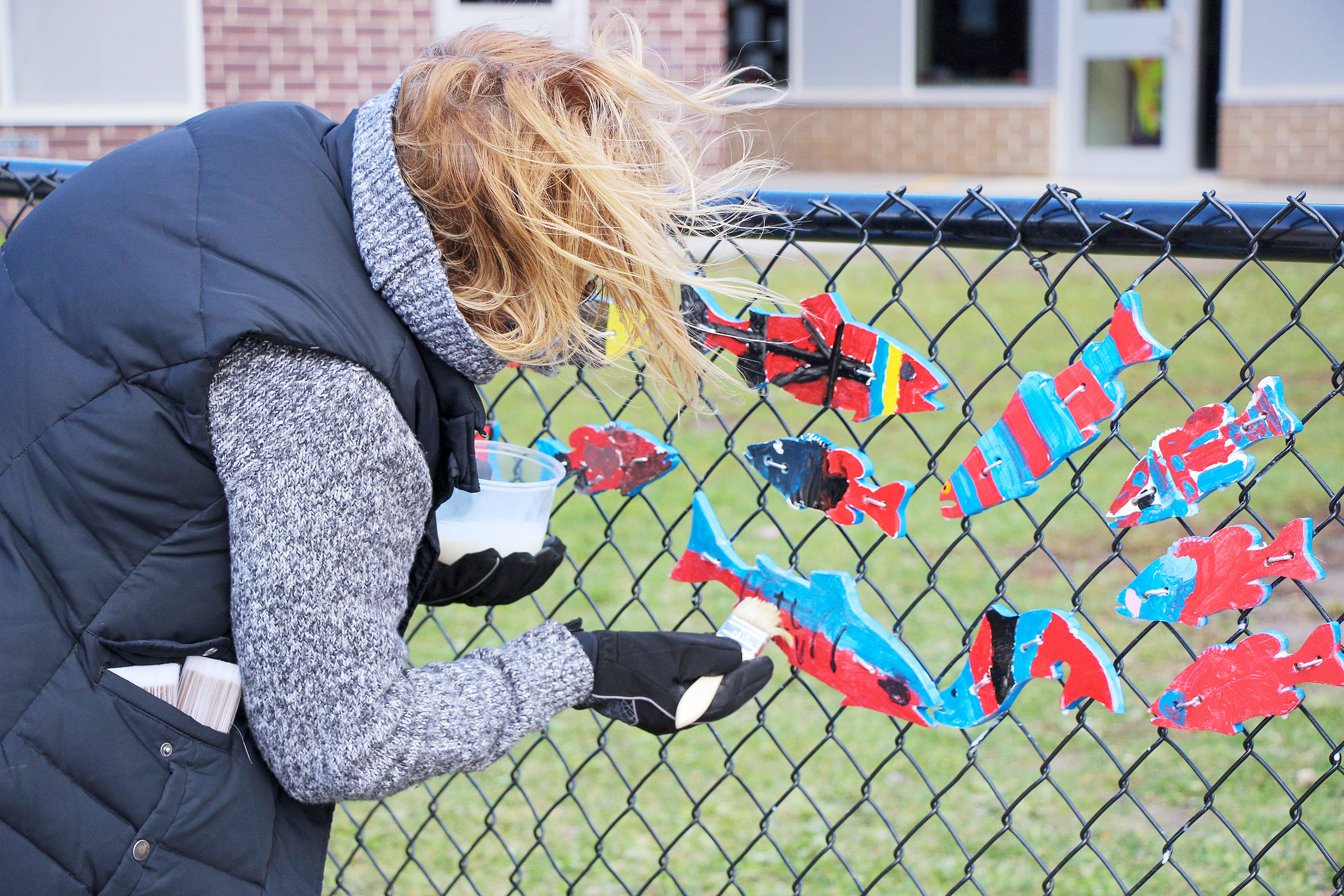 An individual hanging colorful fish cutouts on a chain-link fence outside a school building.
