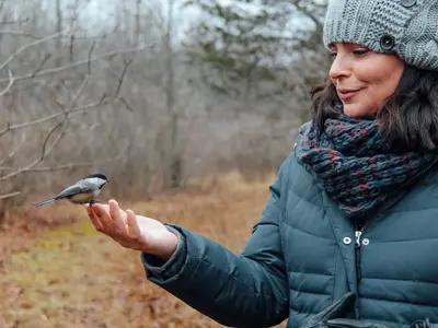 Person in winter clothing holding out hand with a small bird perched on it, standing in a wooded area.