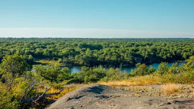 A scenic view of Sheffield Conservation Area featuring a vast expanse of lush greenery surrounding a serene blue lake, with rocky terrain in the foreground under clear blue skies.