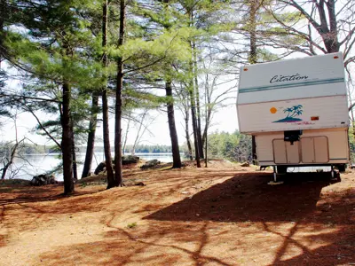 A Clinton travel trailer parked in a wooded lakeside area with tall pine trees and a clear sky.