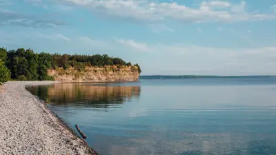 A serene view of a calm lake with a pebbly shore curving towards a cliff covered in greenery under a blue sky with scattered clouds.