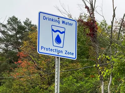 A sign reading "Drinking Water Protection Zone" with a graphic of a water droplet, surrounded by trees and foliage.