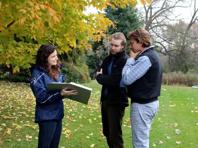 Three individuals having a discussion outdoors with one person holding a clipboard, surrounded by autumn foliage.