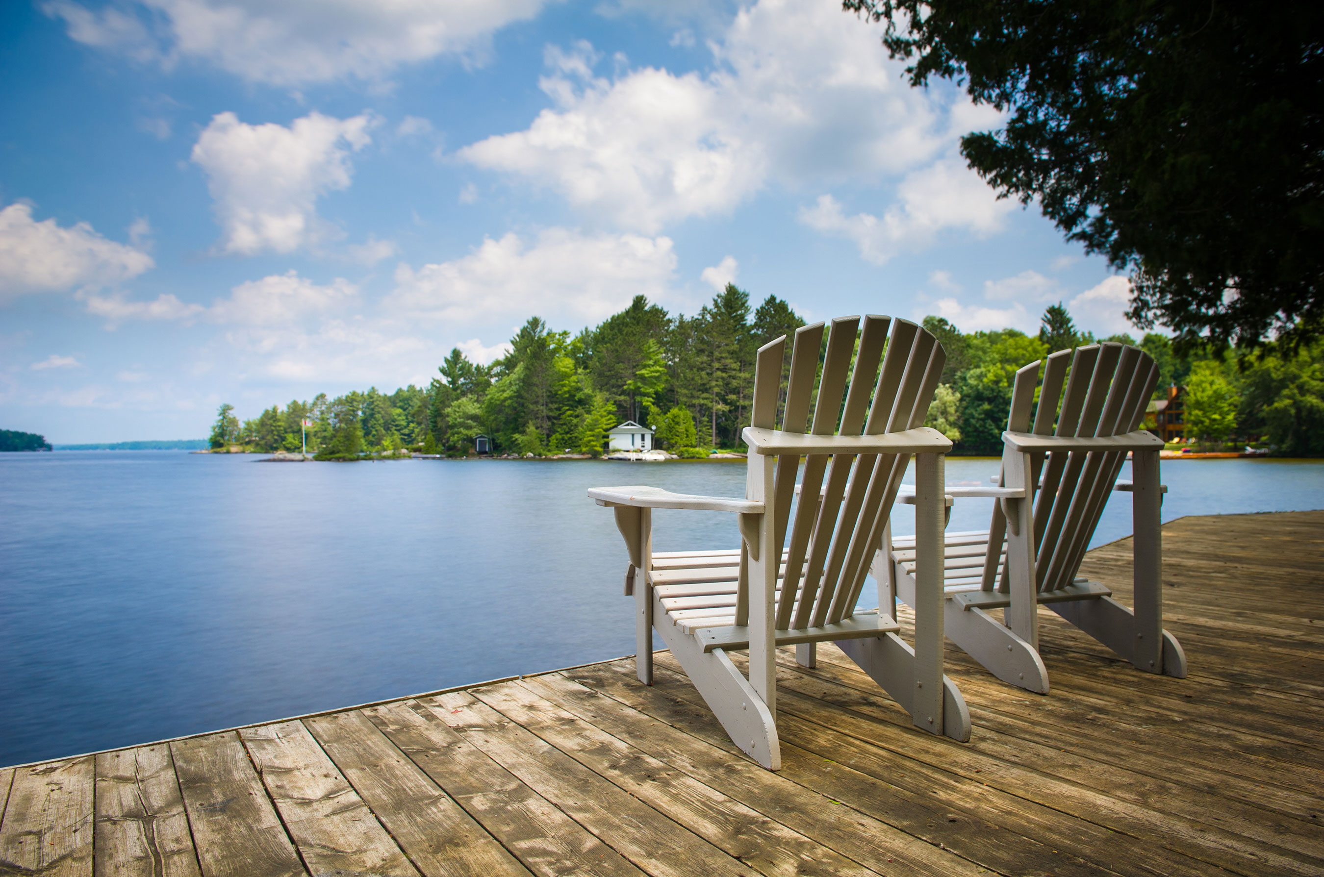 Two Adirondack chairs on a wooden dock overlooking a serene lake with scattered islands and lush trees under a clear blue sky.