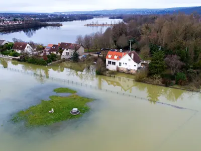 Aerial view of a flooded area with houses near a large body of water, featuring submerged roads and greenery.