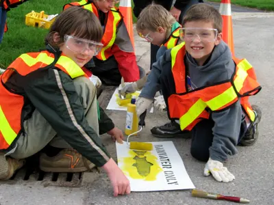 Children wearing safety vests and goggles participate in a paint activity outdoors, working on a 'PAVEMENT ONLY' sign.
