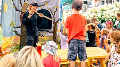 A performer at a public event entertaining a group of children with a storytelling session, using a backdrop that illustrates a tree and props for visual effect.