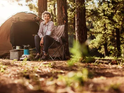 Person sitting on a chair outside a tent, smiling in a forested camping area during the daytime.