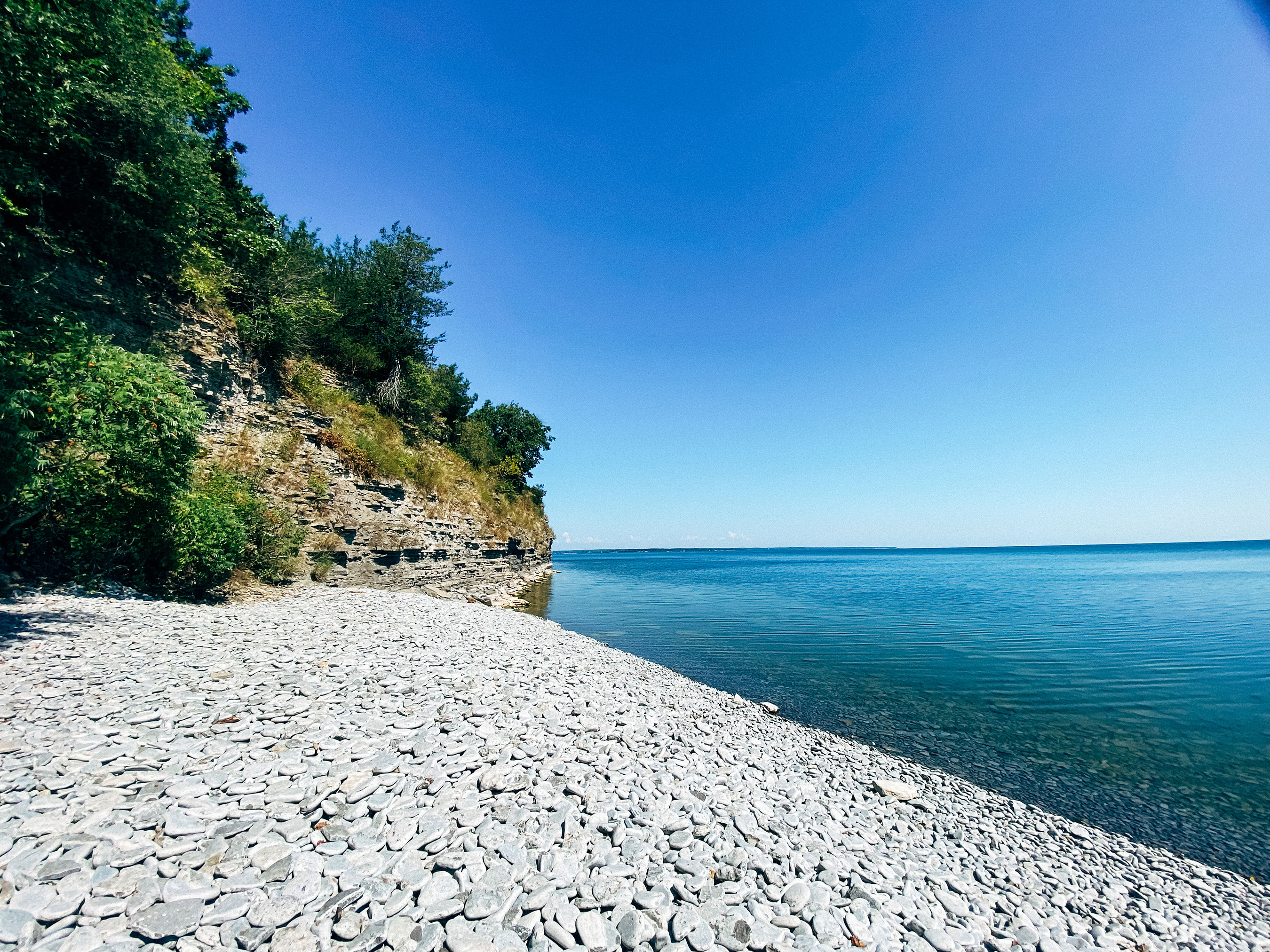 A serene beach with a pebbled shore leading into a clear blue sea, bordered by a lush green cliff on the left under a bright blue sky.