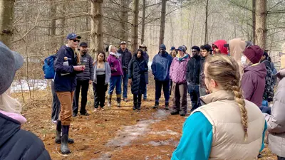 A group of people attentively listening to a speaker during an outdoor excursion in a wooded area.