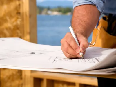 A person in a work apron is drawing on architectural plans at a construction site.