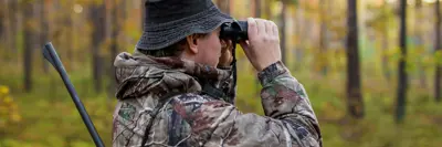 Person in camouflage clothing using binoculars in a forest, holding a shotgun.