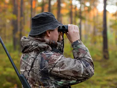 Person in camouflage clothing using binoculars in a forest, holding a shotgun.
