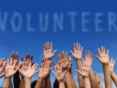 Multiple hands raised towards a clear sky with the word 'VOLUNTEER' written across it in large, white clouds.